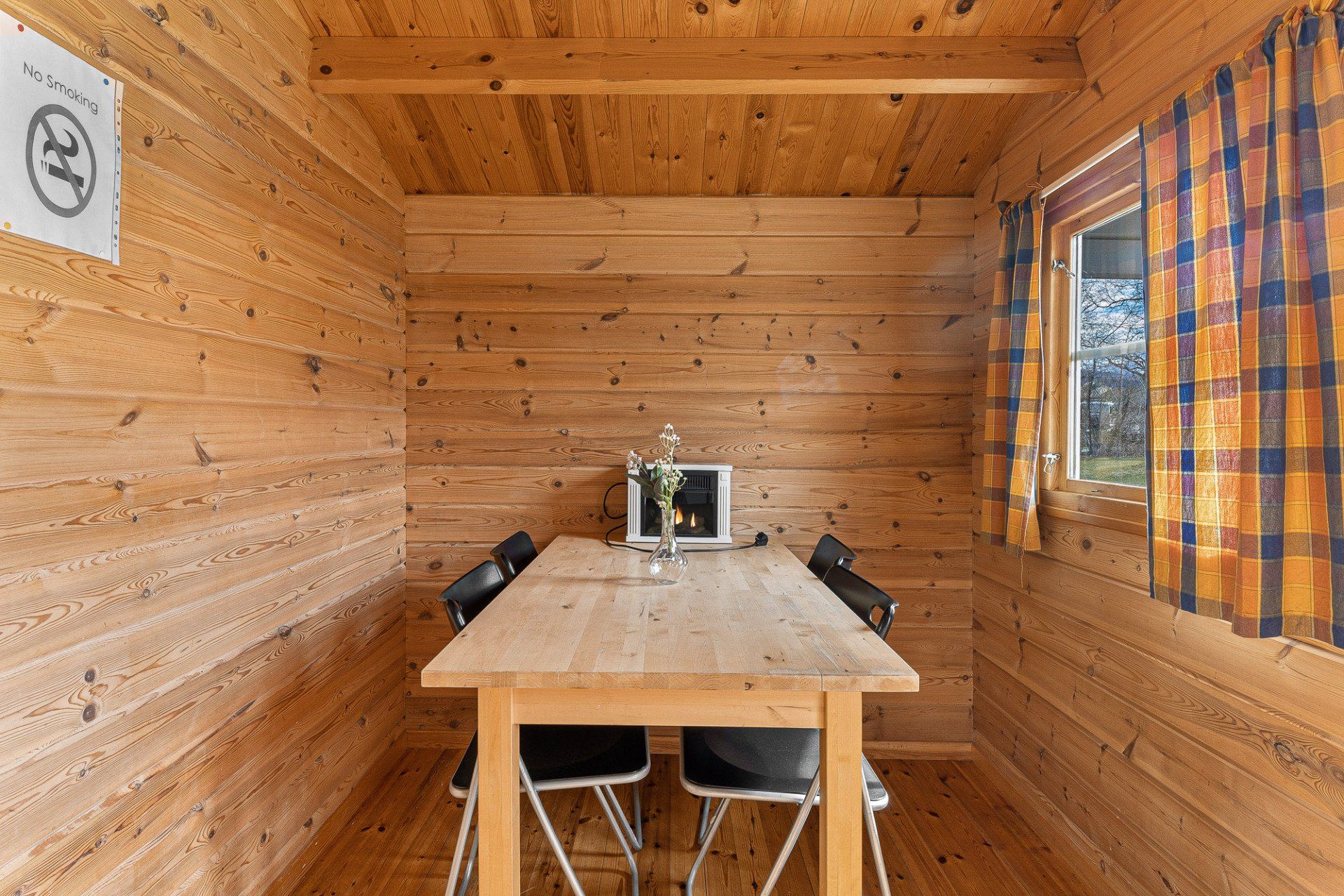 Small wooden cabin interior with a table, chairs, and a window with orange plaid curtains
