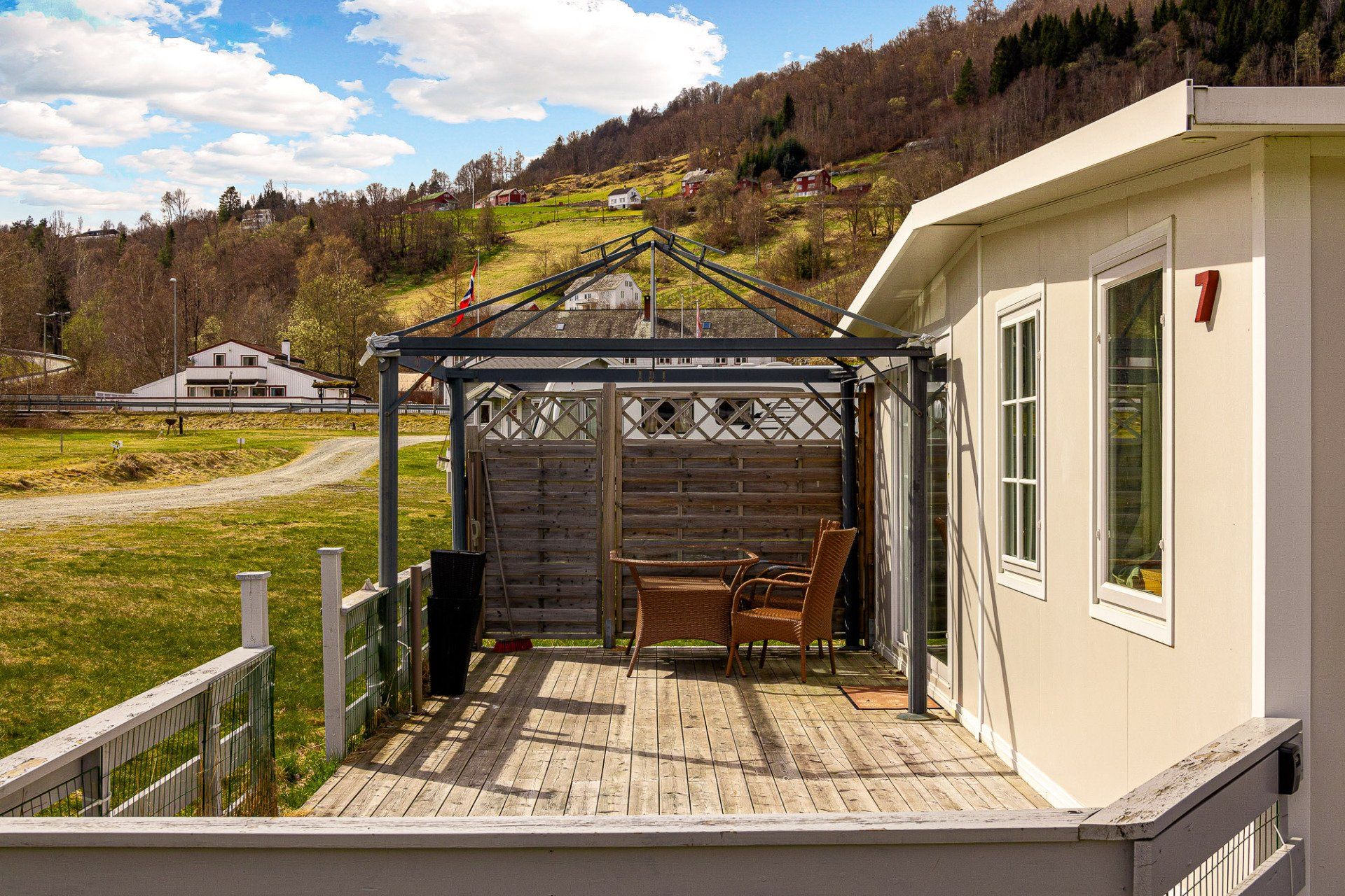 Wooden patio with pergola beside a cream house, overlooking a grassy hillside and cloudy sky.