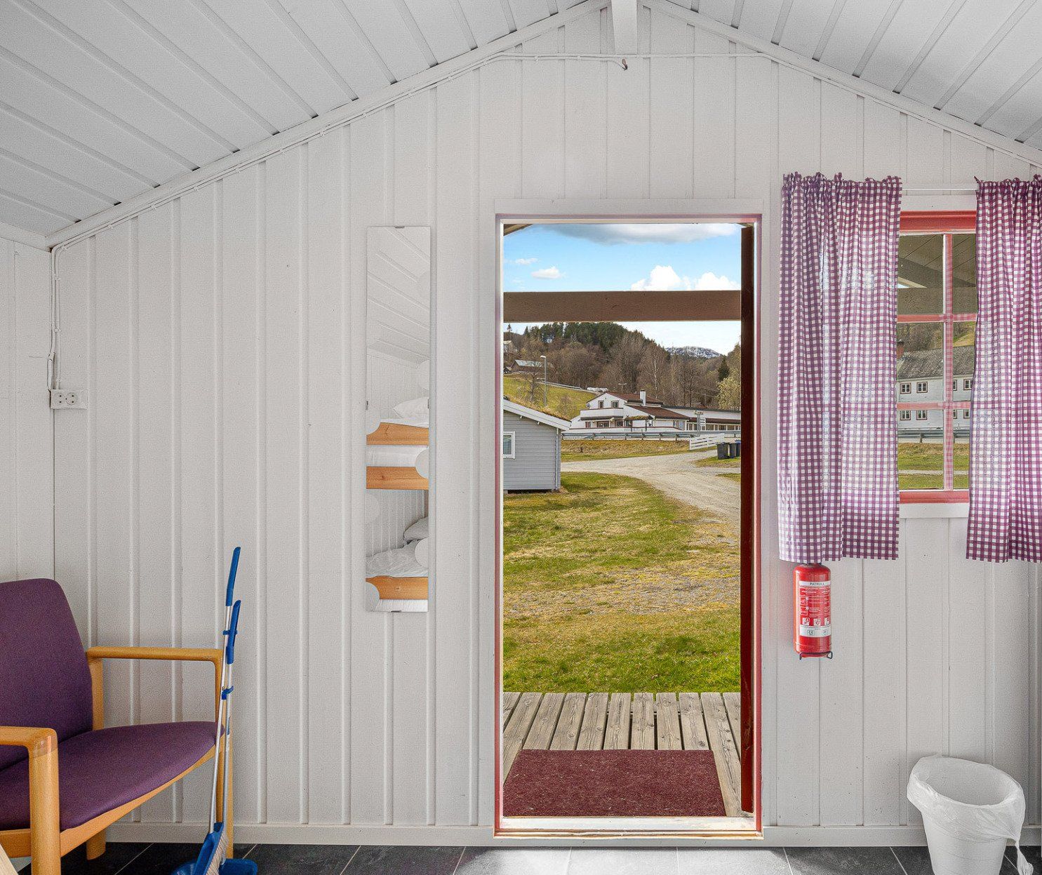 Small white room with a red door opening to a grassy yard and a lake beyond.