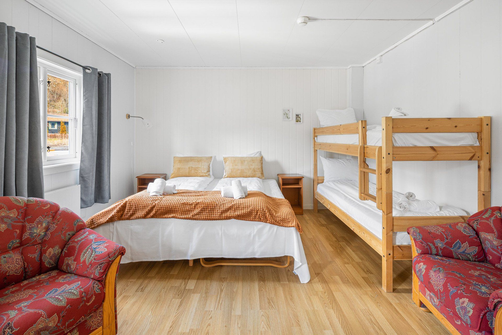 Bright bedroom with a double bed, bunk bed, red armchairs, and a window with gray curtains