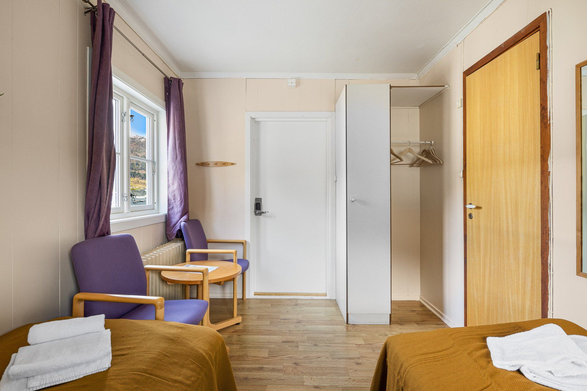 Small bedroom with two beds, purple chair, white wardrobe, and wooden door.