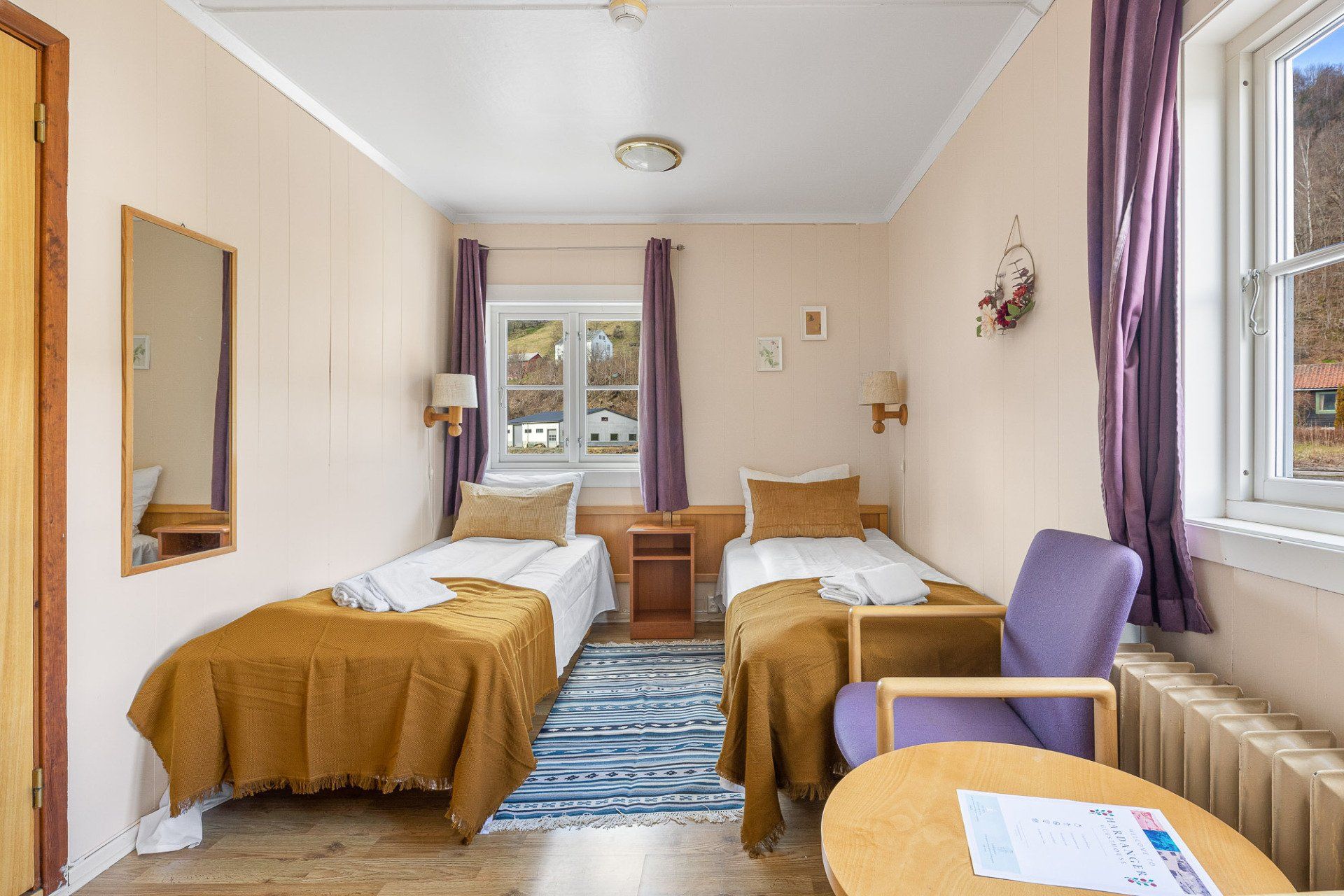 Small bedroom with two twin beds, blue rug, purple curtains, and a chair by the window.