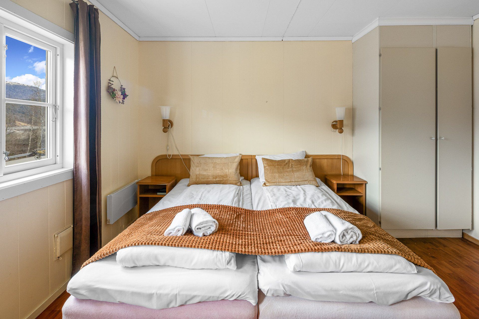 Bright bedroom with a double bed, wooden nightstands, white wardrobe, and a large window showing mountains.