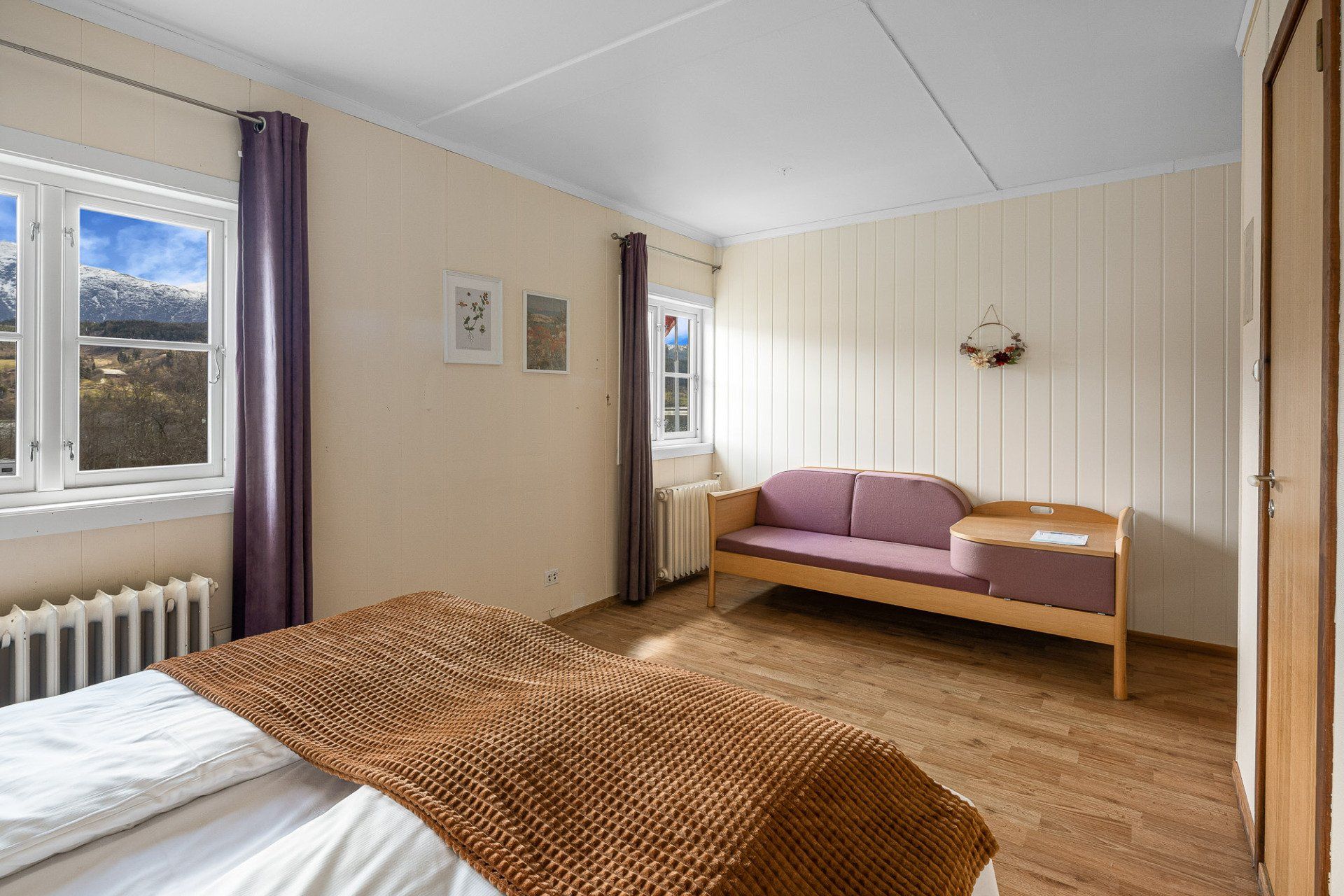 Bright bedroom with bed, purple sofa, wood floor, and windows with curtains
