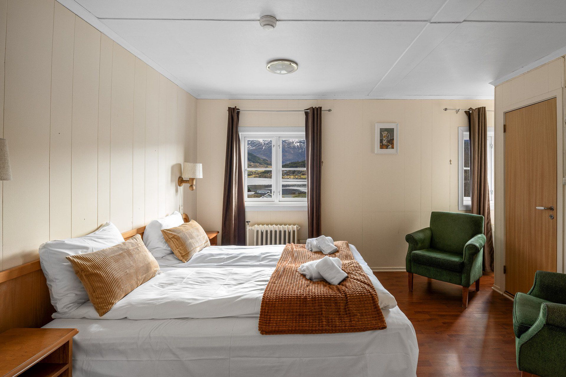 Cozy bedroom with a large bed, green chairs, wood paneling, and a window with a view
