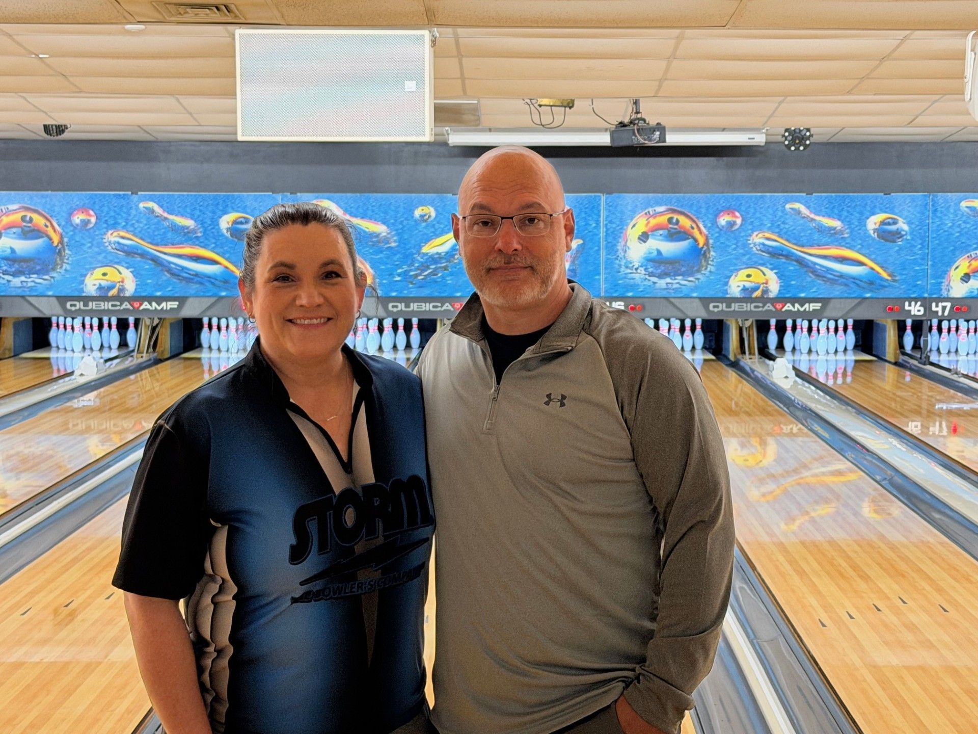 Two people standing in a bowling alley, smiling in front of the lanes.