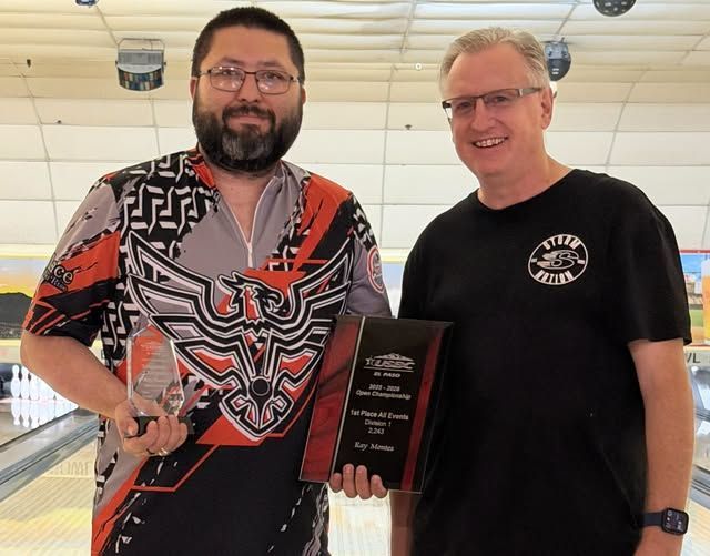 Two men at a bowling alley holding an award plaque and a trophy, smiling at the camera.