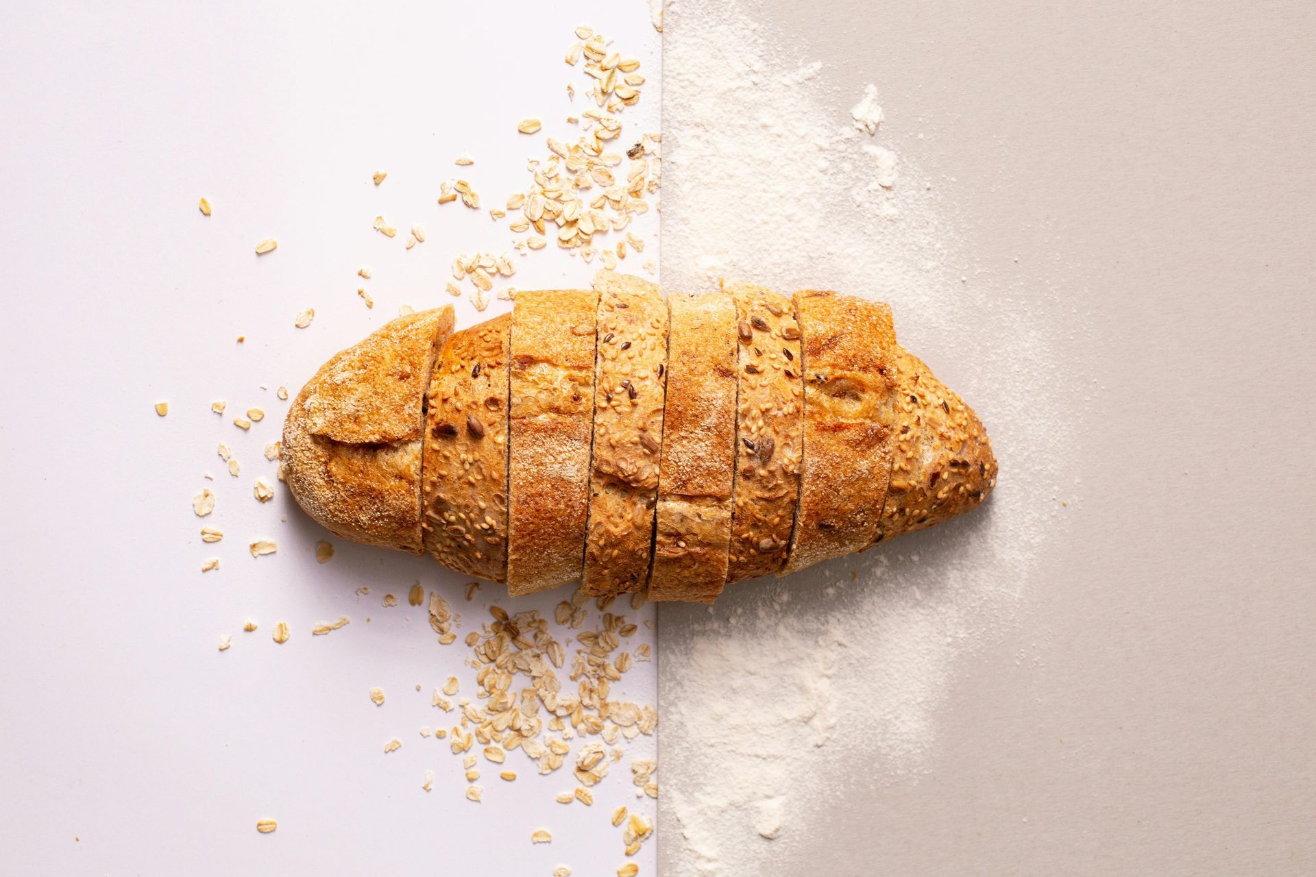 Sliced loaf of bread on a surface split into white and gray sides, with oat grains and flour scattered — All Body Holistic Health in Thuringowa Central, QLD
