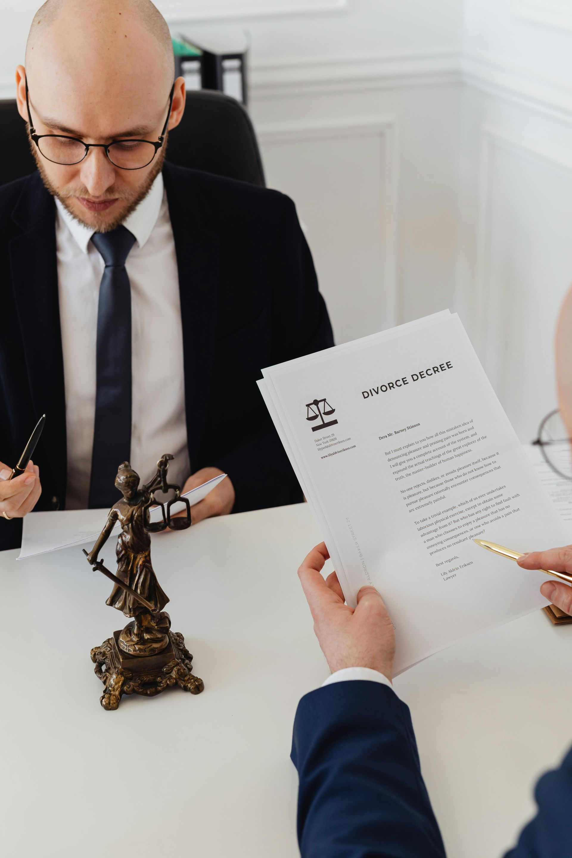 Dos personas trajeadas revisan documentos en un escritorio. Sobre él hay una estatua de la Dama de la Justicia.