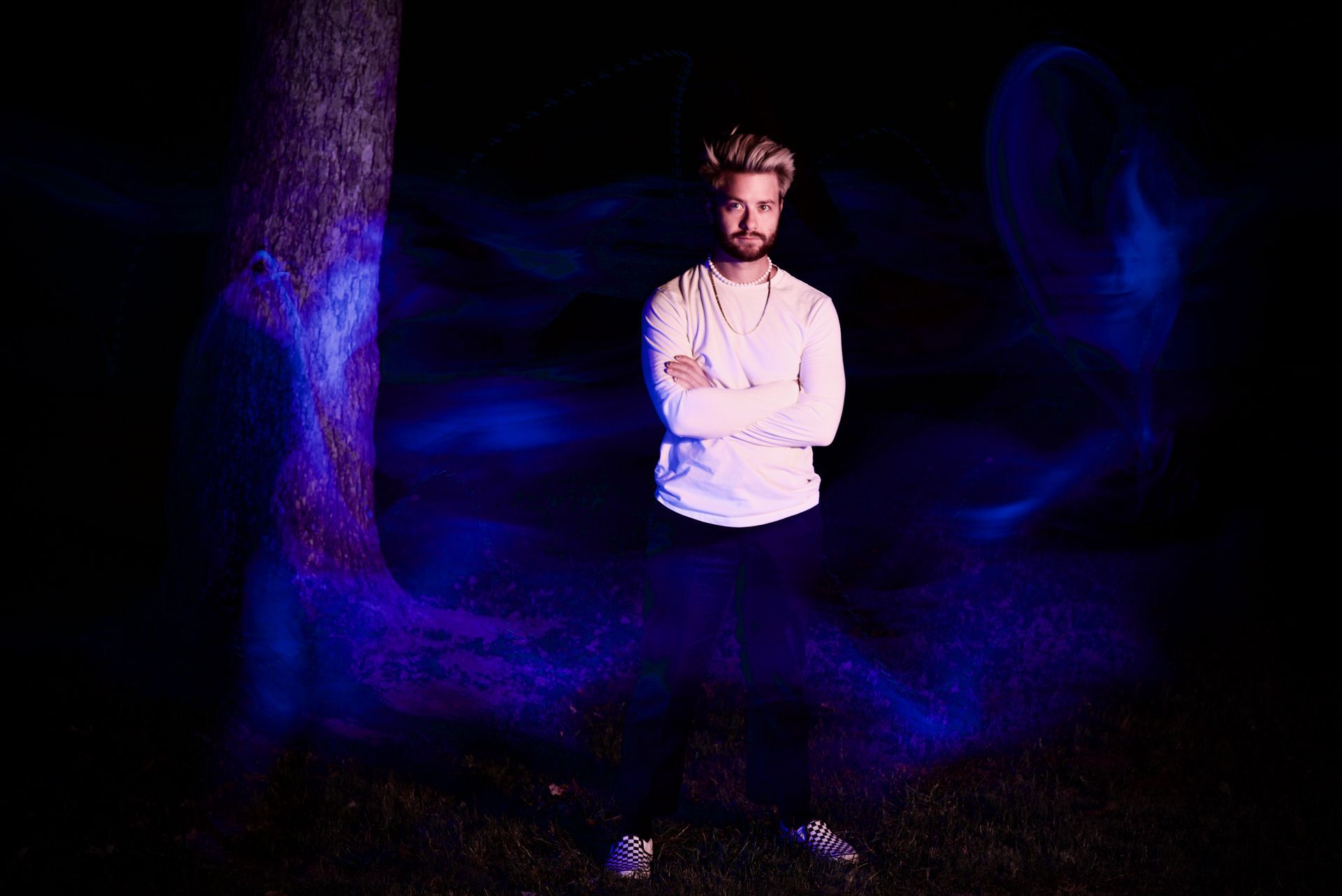 A man is standing in front of a tree in the dark with his arms crossed.
