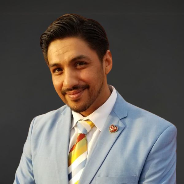 Man in a light blue suit with a striped tie, smiling against a dark background
