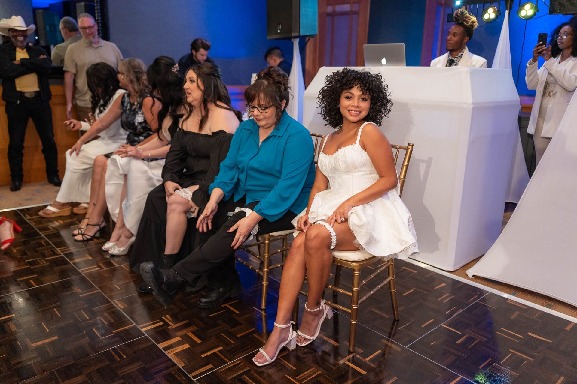 Bride sits on a chair, garter on her thigh, surrounded by guests at an event. DJ booth behind.