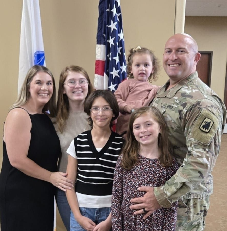 Group posing indoors in front of flags, with one man in military uniform holding a child.