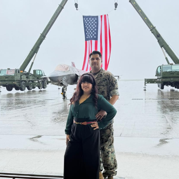 Couple posing on a wet airfield between military vehicles, with a large U.S. flag in the background.