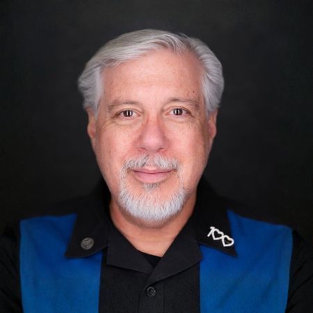 Man with gray hair and beard, wearing black shirt and blue collar, smiling.