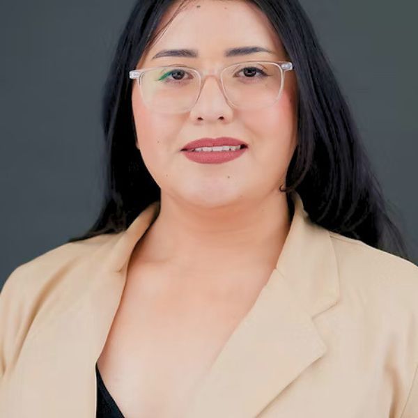 Maggie Millan, wearing a tan blazer, posing in front of a stadium seating background.