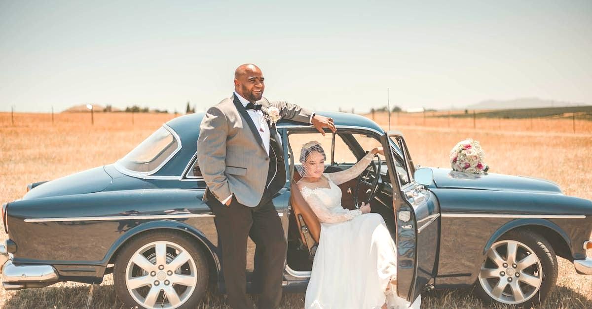 A bride and groom are standing next to a blue car in a field.