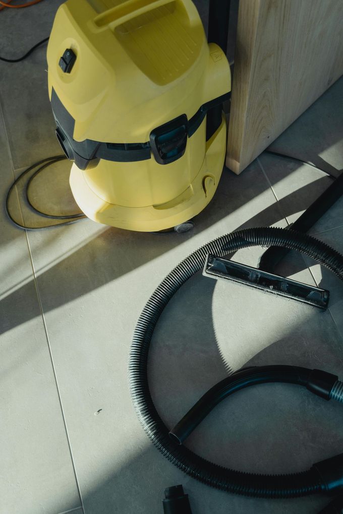 Yellow vacuum cleaner on a sunlit floor beside a wall, with black hose coiled nearby.
