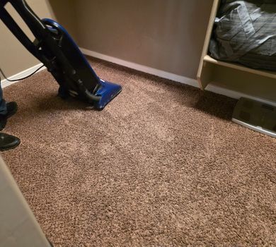 Vacuum cleaner on beige carpet in a small room corner with shelving and a wall vent