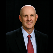 Studio portrait of a bald man in a dark suit, light blue shirt, and red tie against a black background
