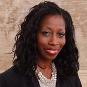Smiling woman with long curly black hair, wearing a black blazer and pearl necklace against a beige background