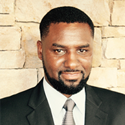 Portrait of a man in a black suit and tie against a stone wall, smiling softly