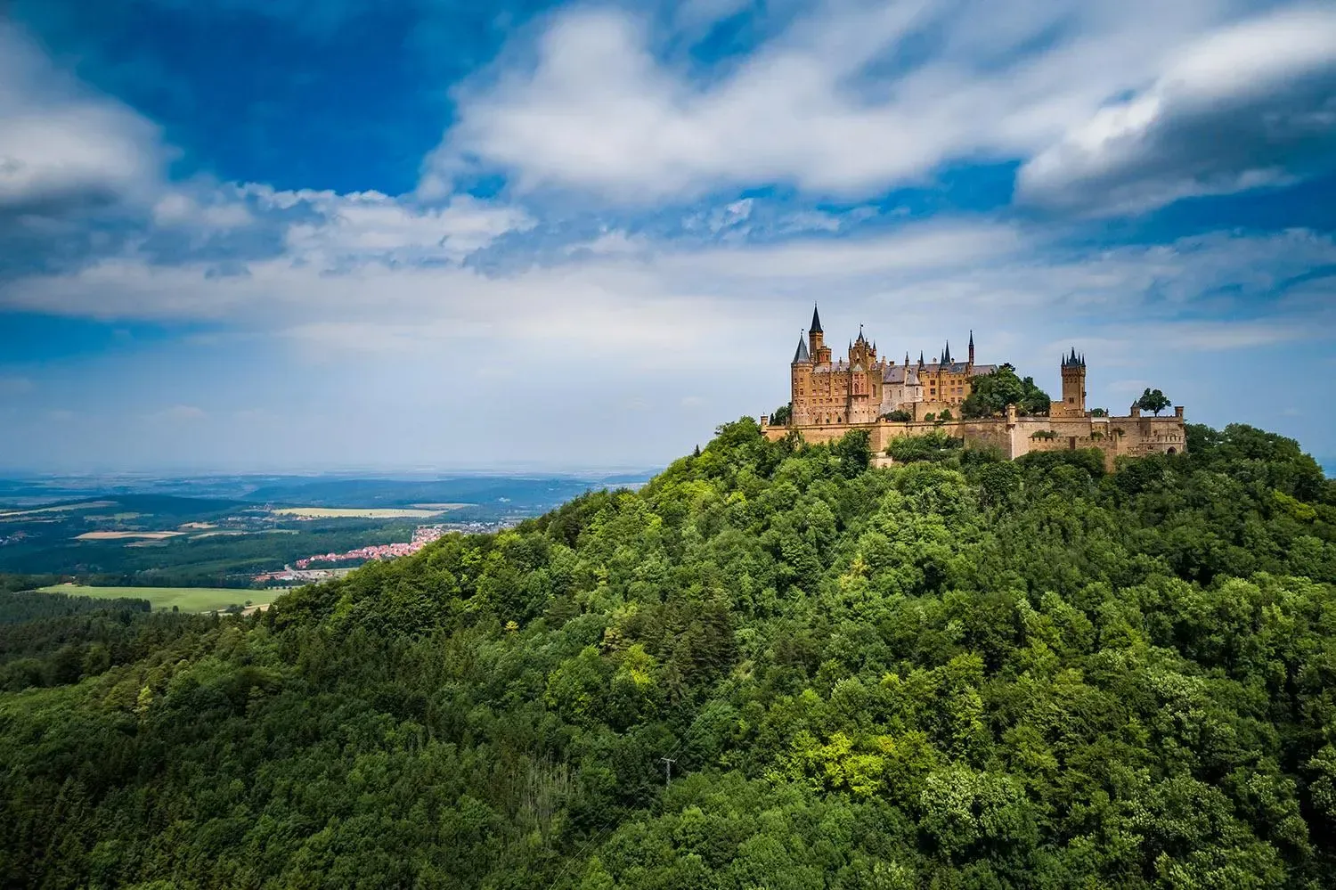 Castle atop a green, forested hill against a blue sky with clouds.