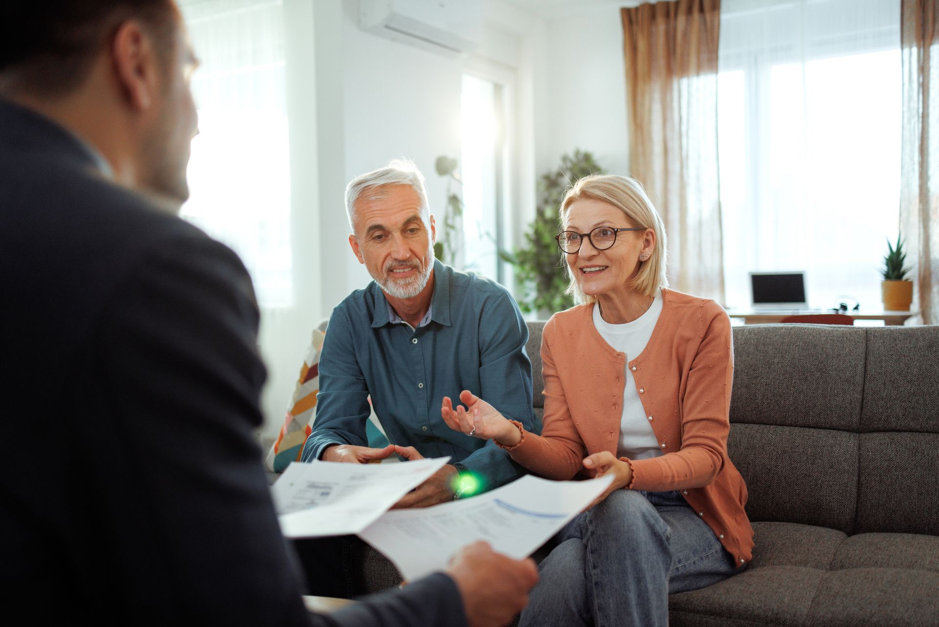 Man in suit reviewing documents with a senior couple in a living room. The woman gestures while speaking.