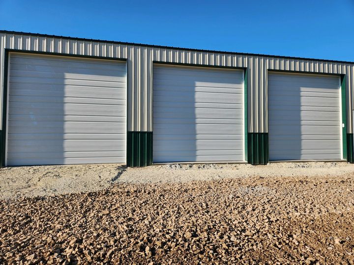 Three closed metal garage doors on a corrugated building with a gravel foreground under a blue sky