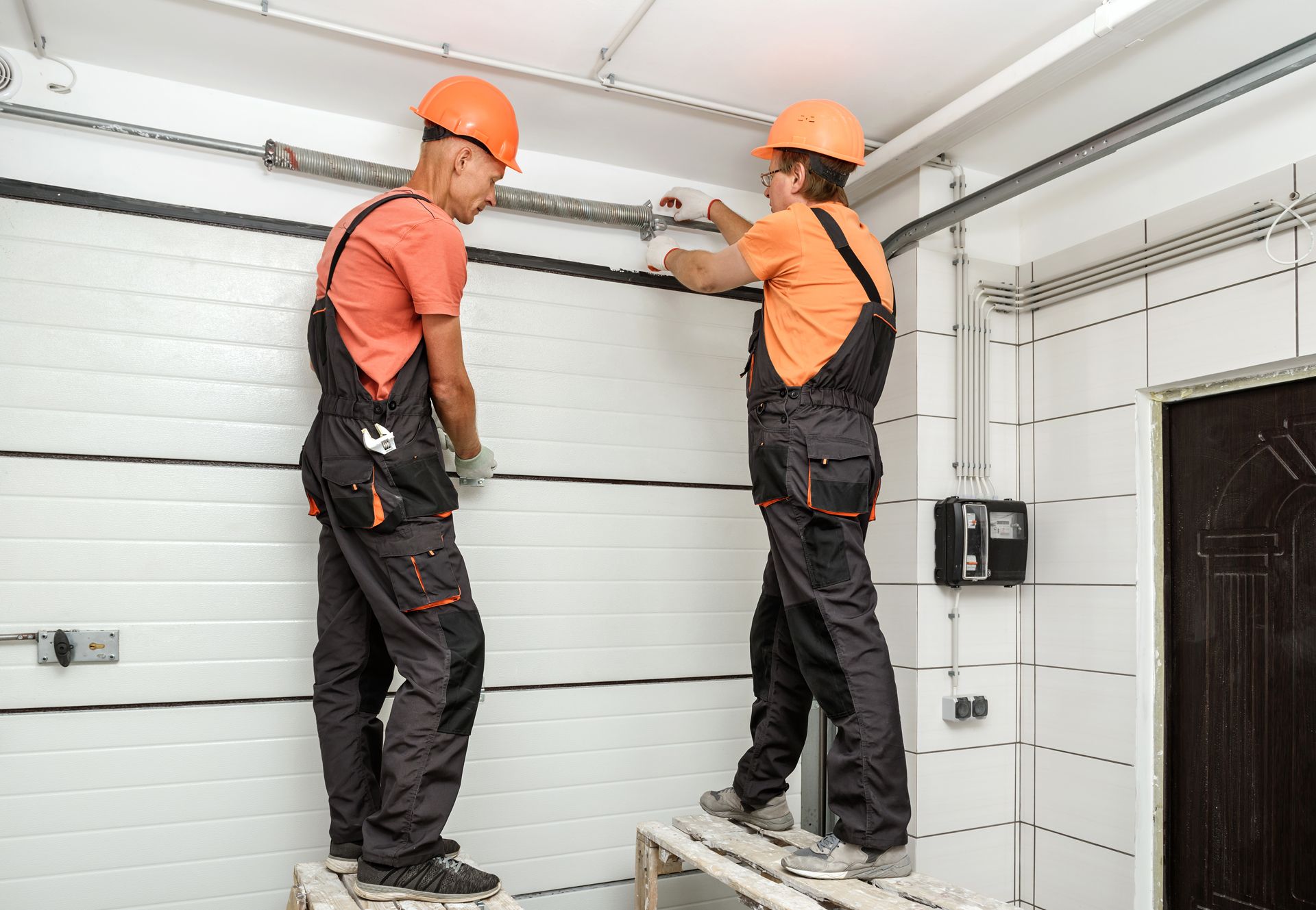 Two workers in orange hard hats adjusting a large white garage door panel indoors