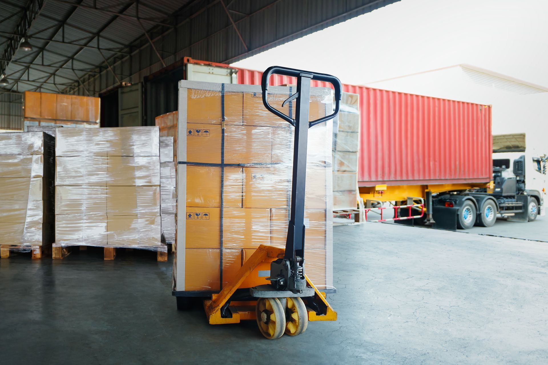 Warehouse forklift moving stacked pallets near a red shipping container