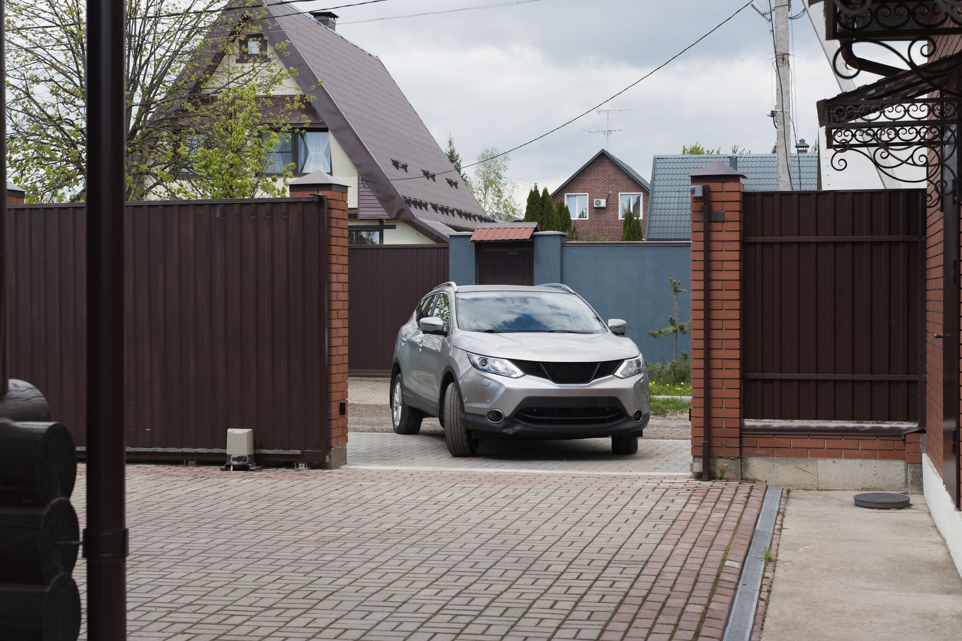 Silver SUV parked in a paved driveway between open brown gates and brick pillars at a residential home