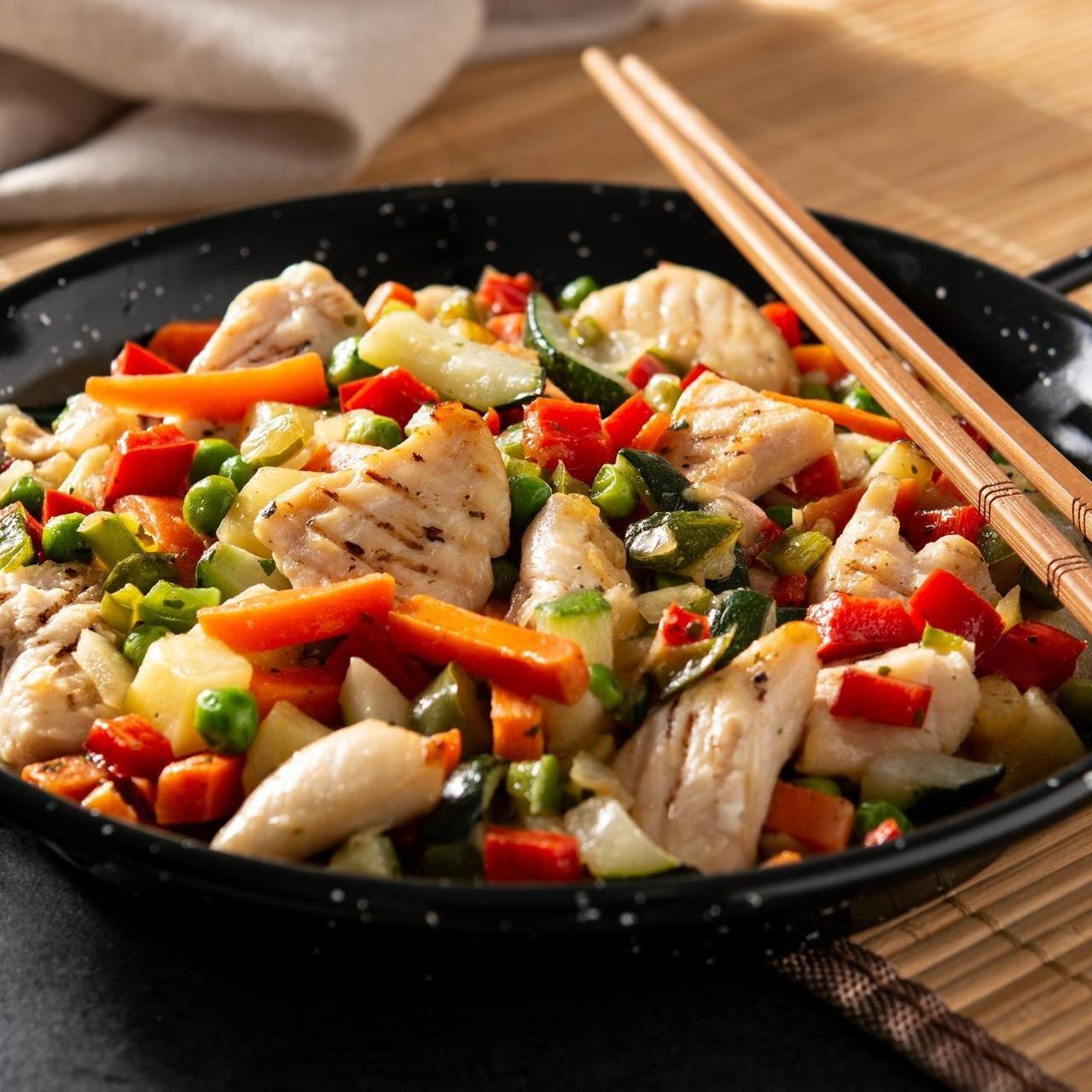 A black pan filled with chicken and vegetables next to chopsticks
