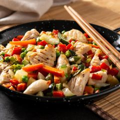 A black pan filled with chicken and vegetables next to chopsticks