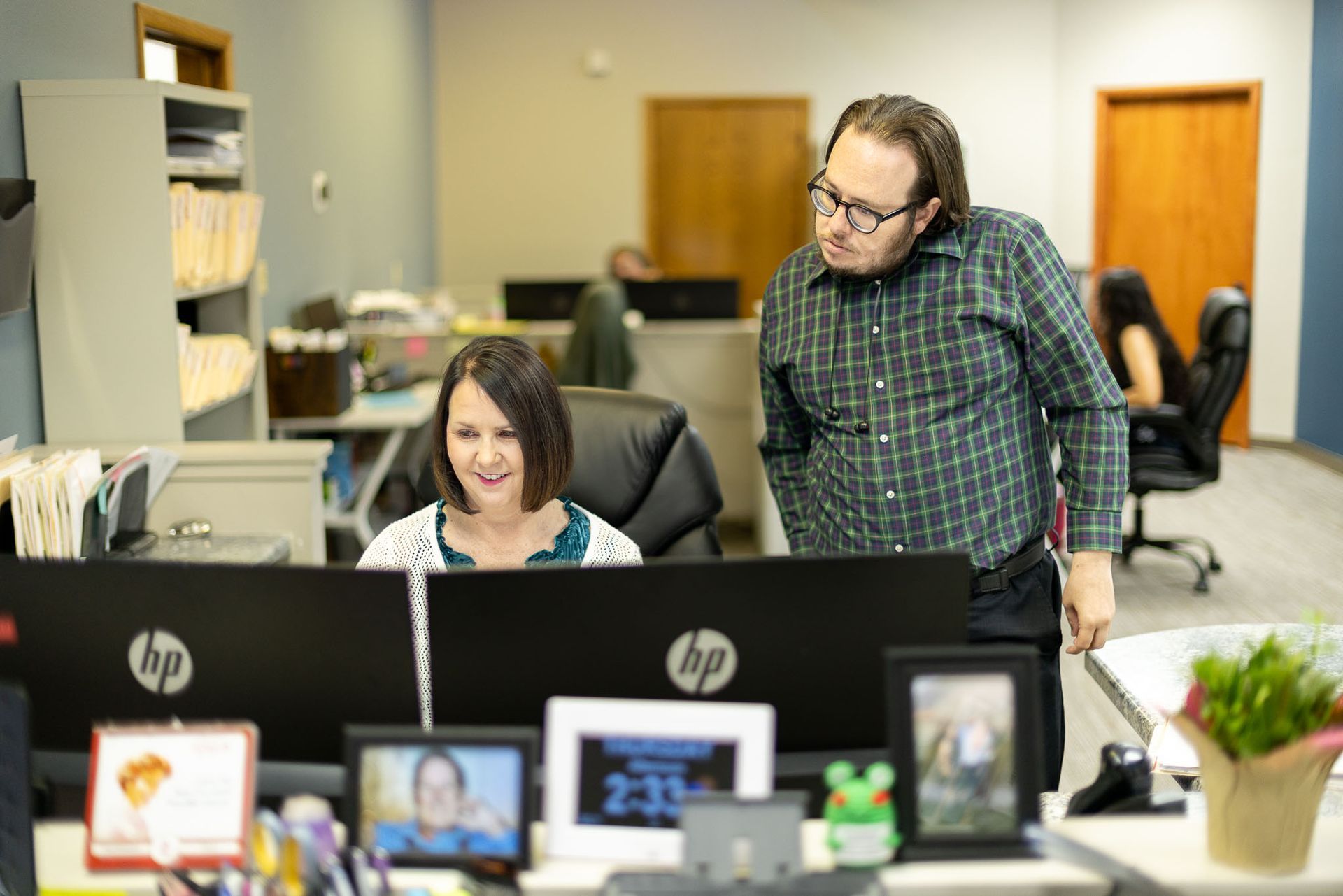 Two coworkers at desks in an office, one seated and smiling at a computer while the other stands beside her.