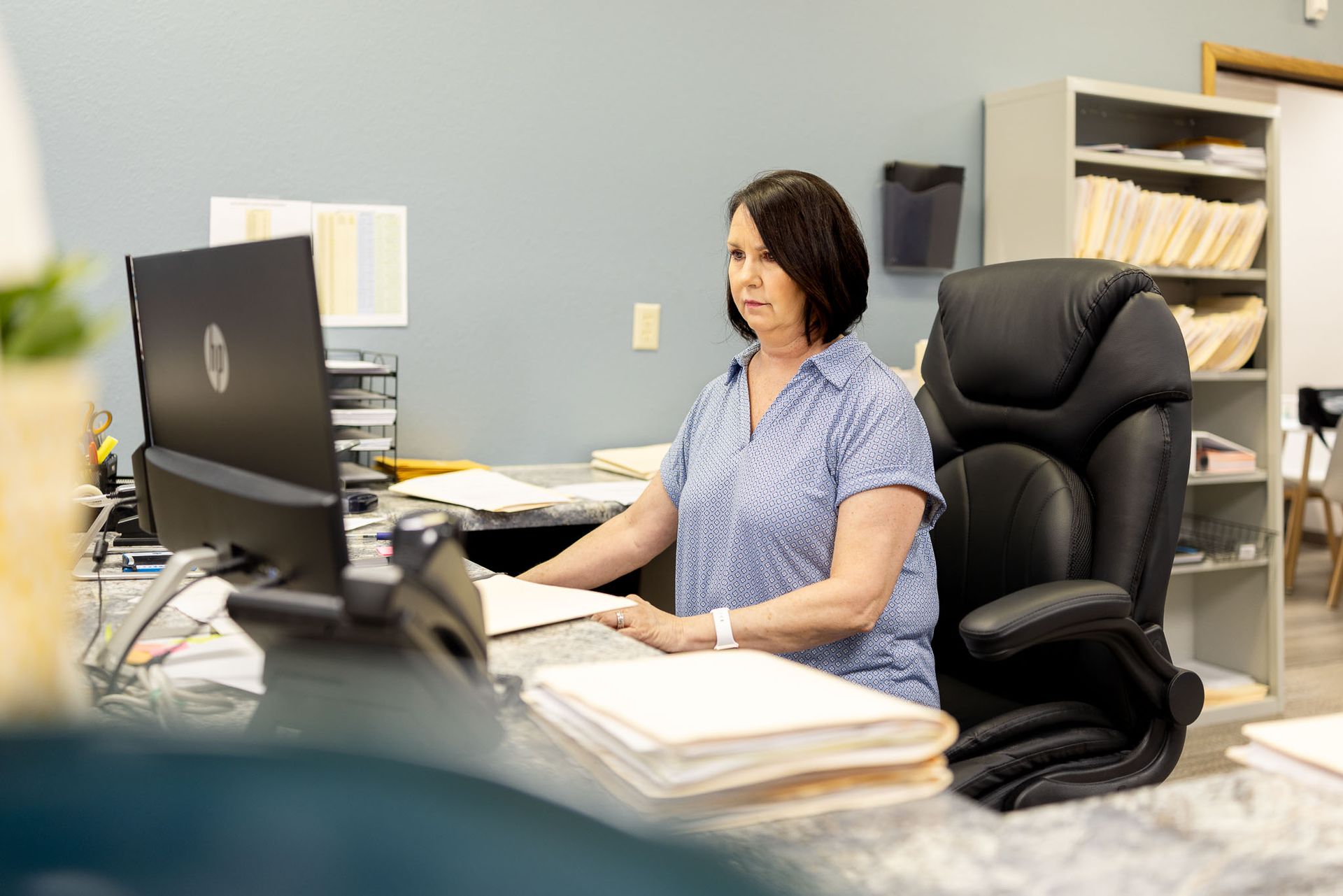 Woman working at a desk in an office, seated beside a computer with papers and files nearby.