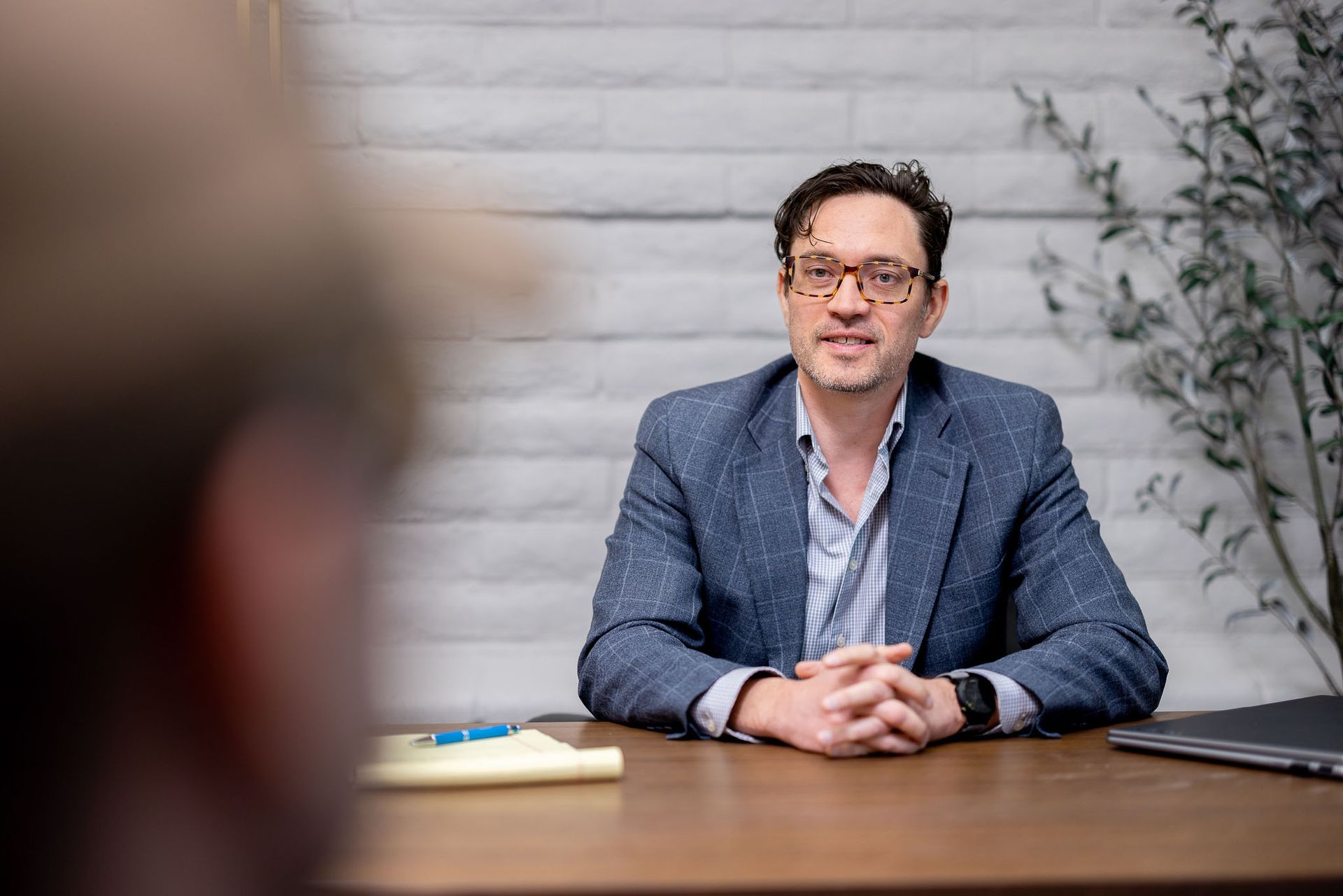 Ken in a blue blazer sits at a table in an office, facing forward with hands clasped.
