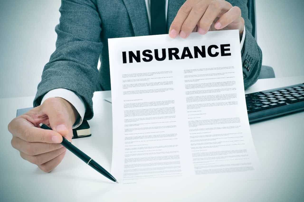 Person holding a pen beside an insurance document on a desk, with laptop in the background