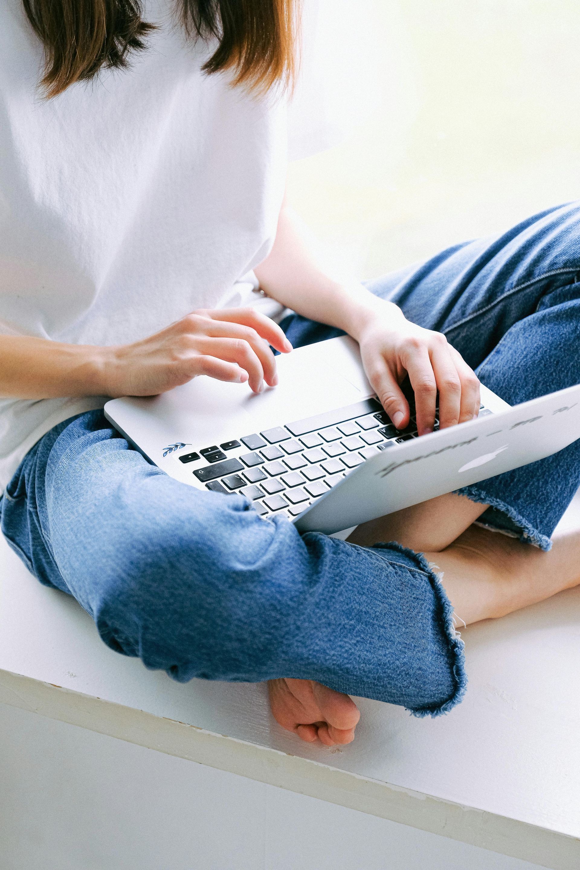 Person sitting cross-legged on a bed using a laptop, wearing jeans and a white shirt.