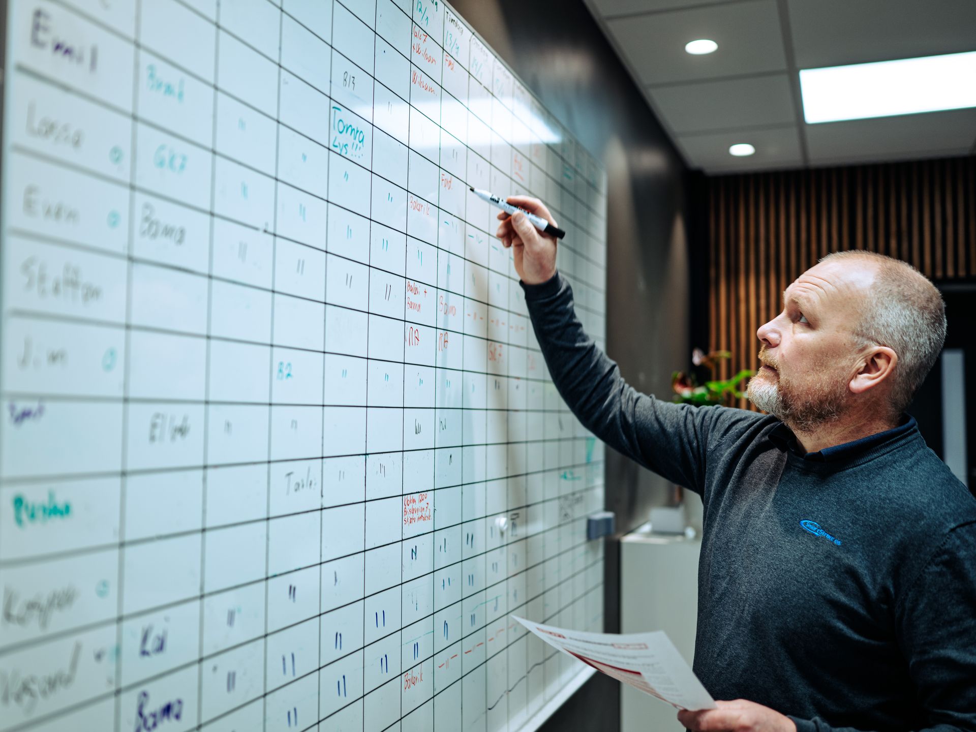 En person står på et kontor og skriver på et stort tavlenett mens han holder et papirdokument.