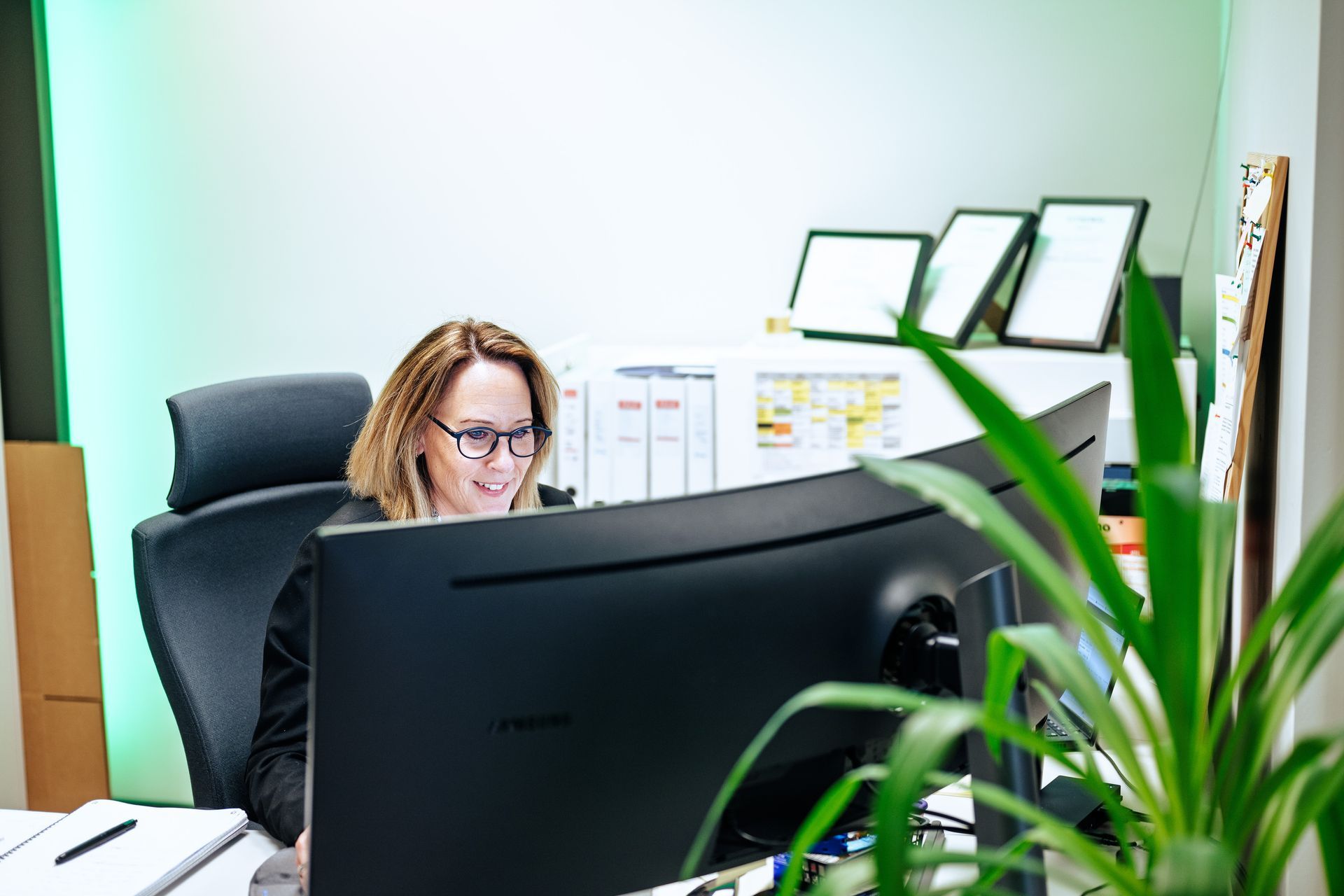En smilende person sitter ved et skrivebord på et kontor og arbeider bak en stor, buet dataskjerm.