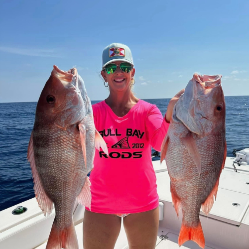 A woman wearing a full bay rods shirt is holding two fish