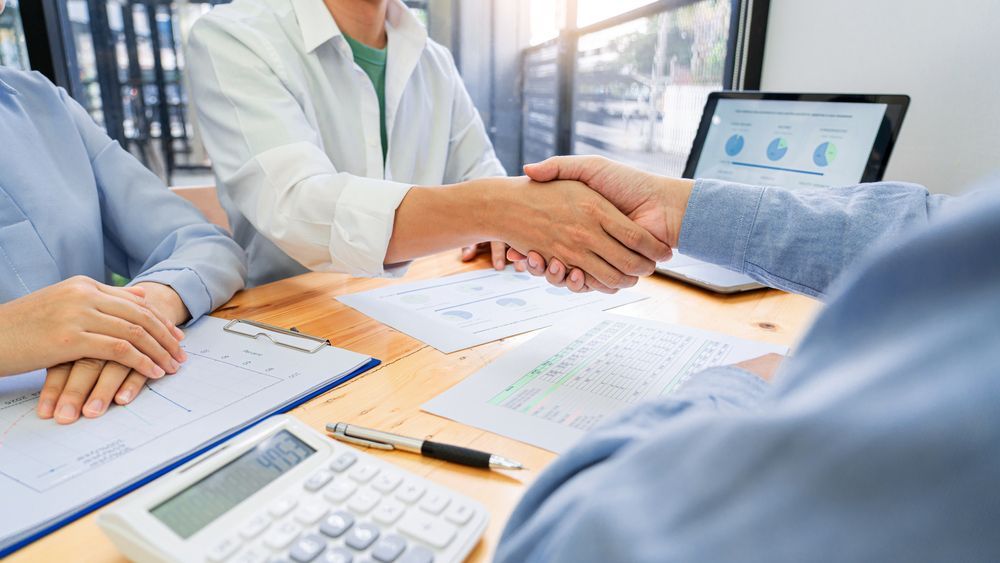 Colleagues in a meeting shaking hands over documents and charts on a conference table