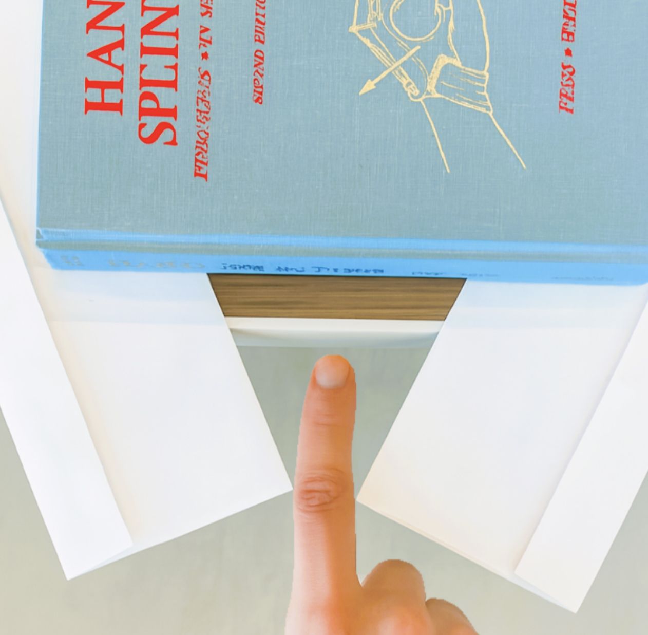 Finger holding open a blue book, showing a triangular white paper shape underneath.