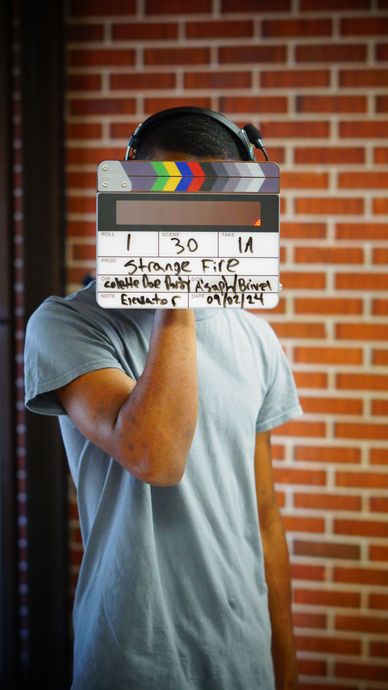 Person holding a clapperboard in front of a brick wall, face hidden behind it.