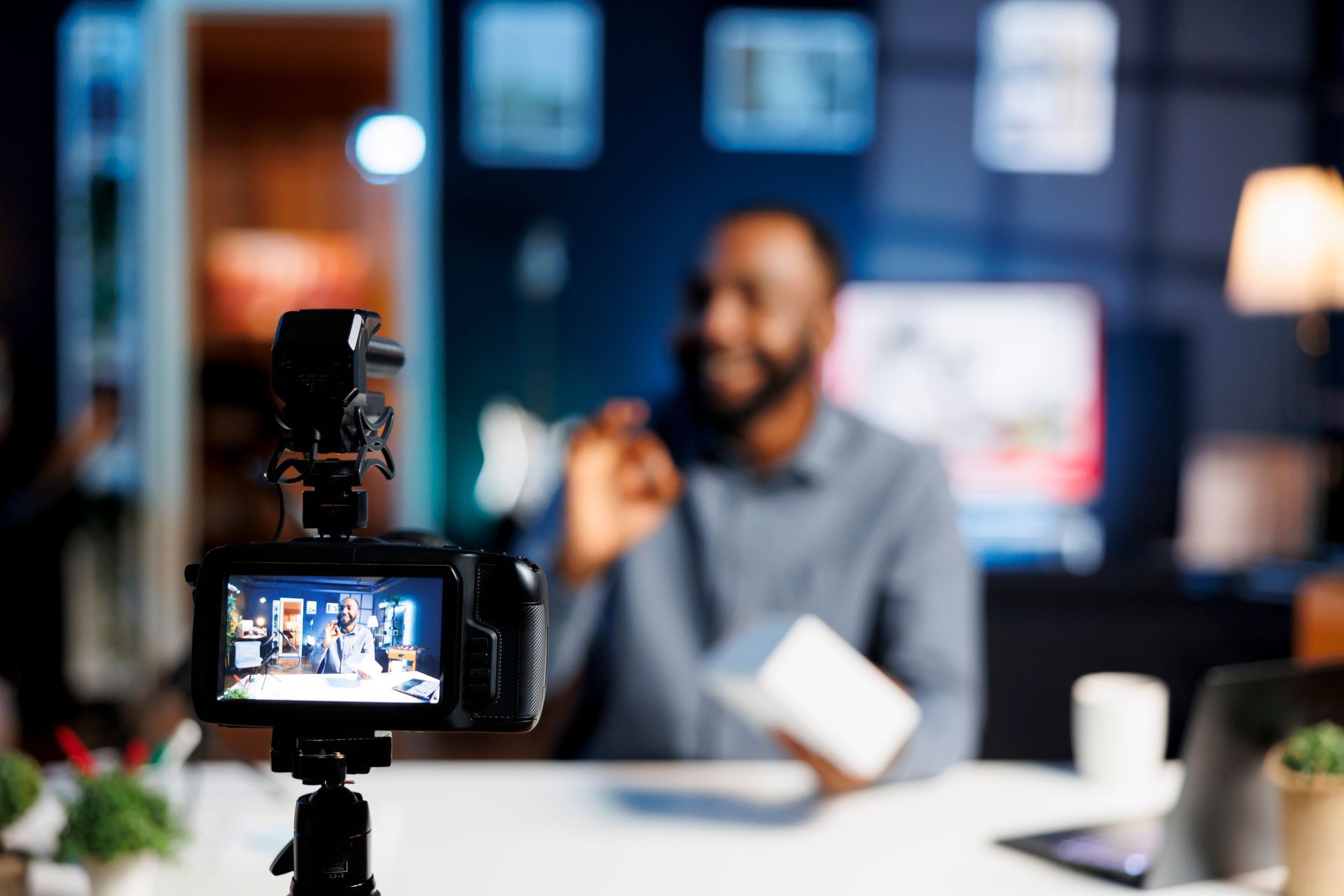 Camera filming a smiling speaker at a desk in a blurred studio setting