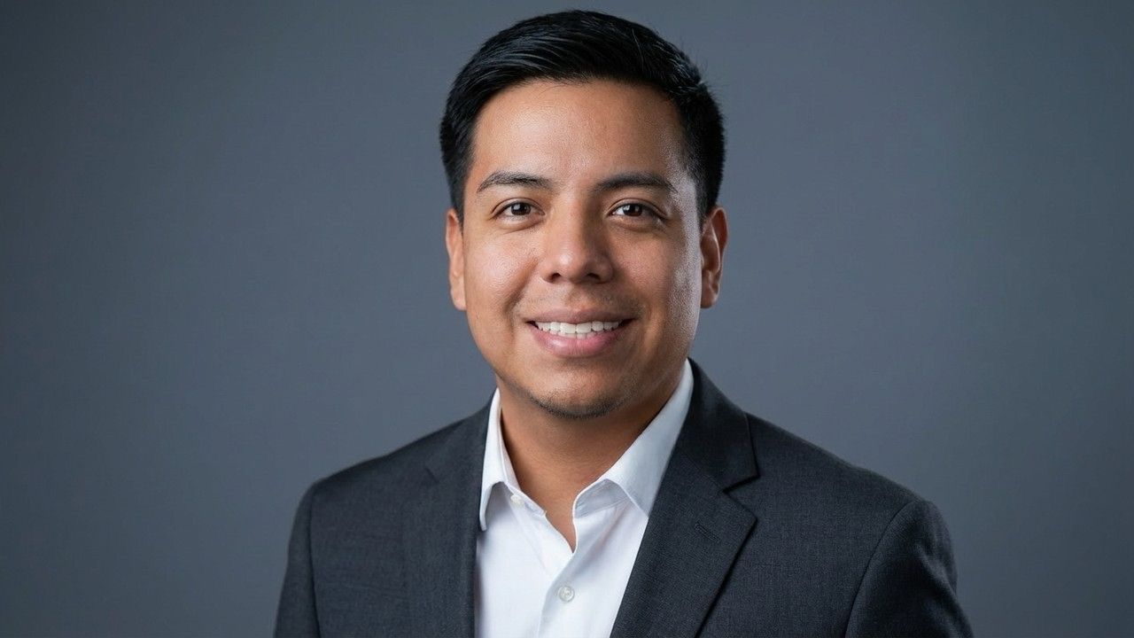 Man in a grey suit and white shirt, smiling, standing against a light grey background.