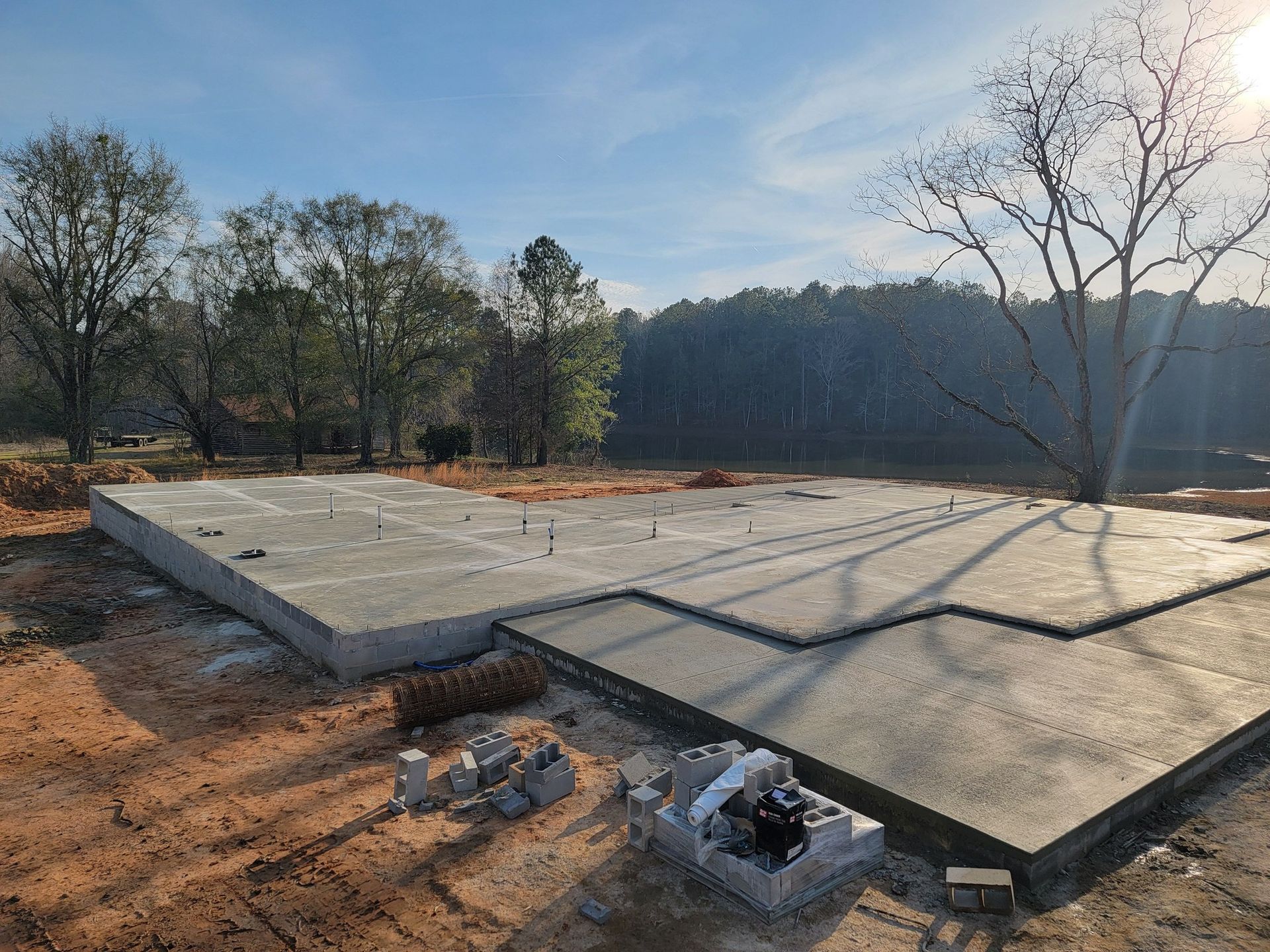 Concrete foundation for a building in a dirt lot, surrounded by trees under a blue sky.