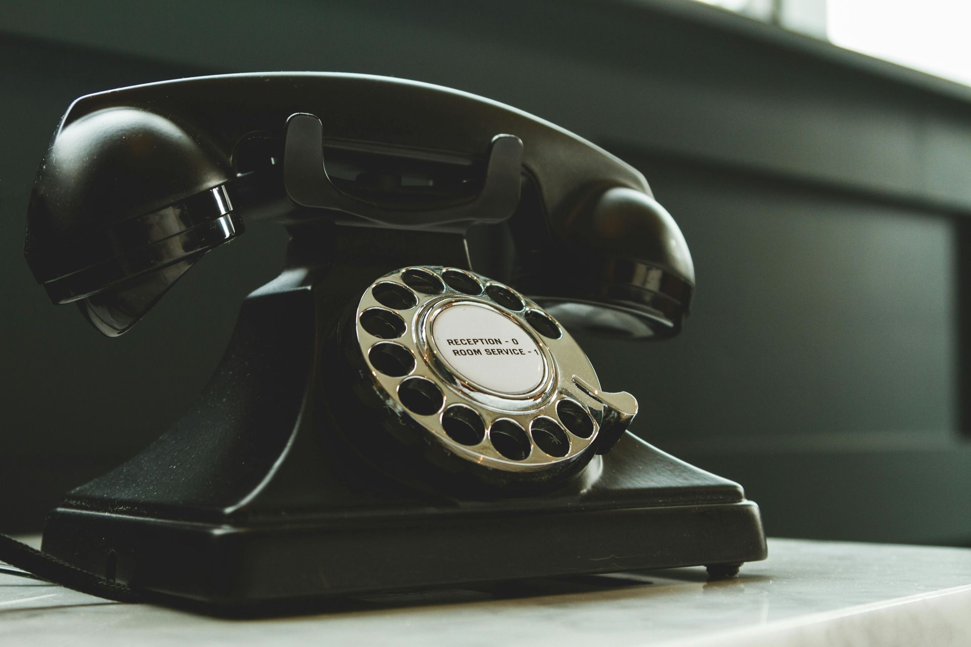 A classic black rotary telephone sits on a light surface against a dark, blurred background.