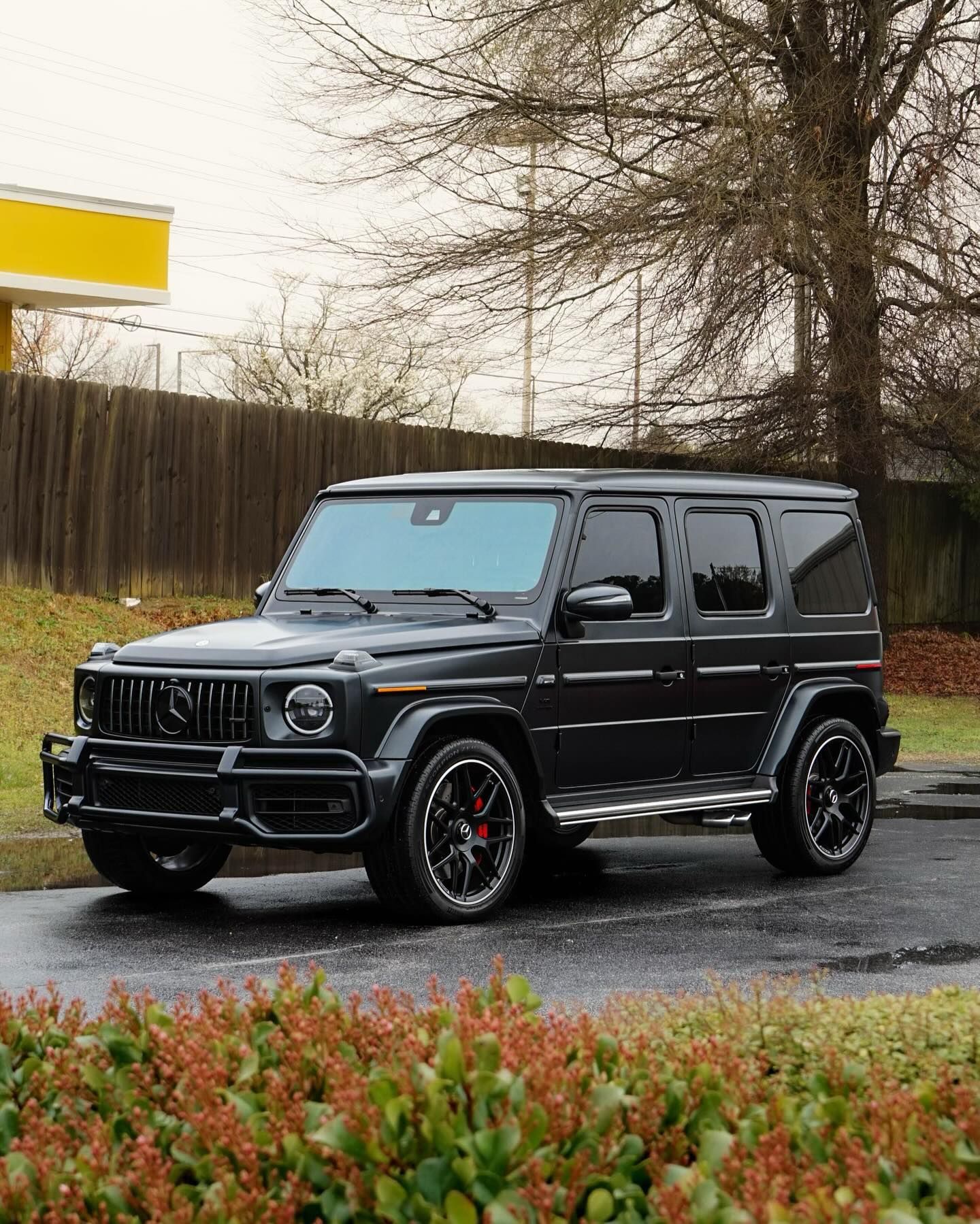 Dark gray Mercedes-Benz G-Wagon parked on wet pavement near shrubs and a wooden fence.
